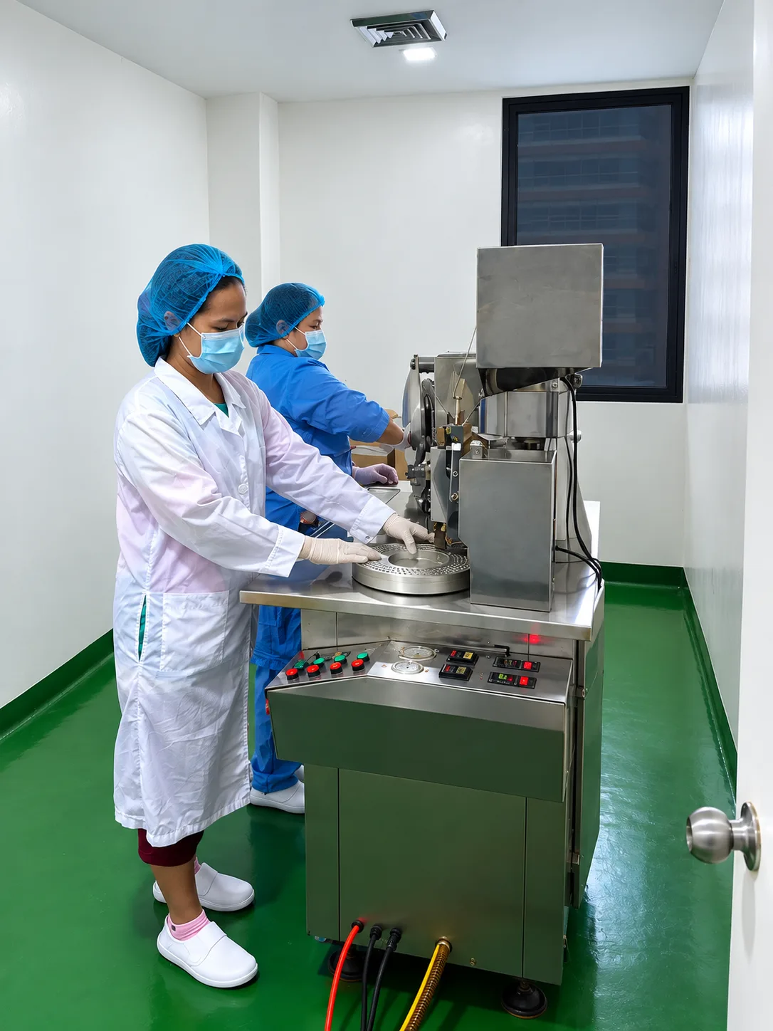Encapsulation Room 2: two workers wearing masks, hair coverings, and gloves operating stainless-steel equipment with circular tooling.